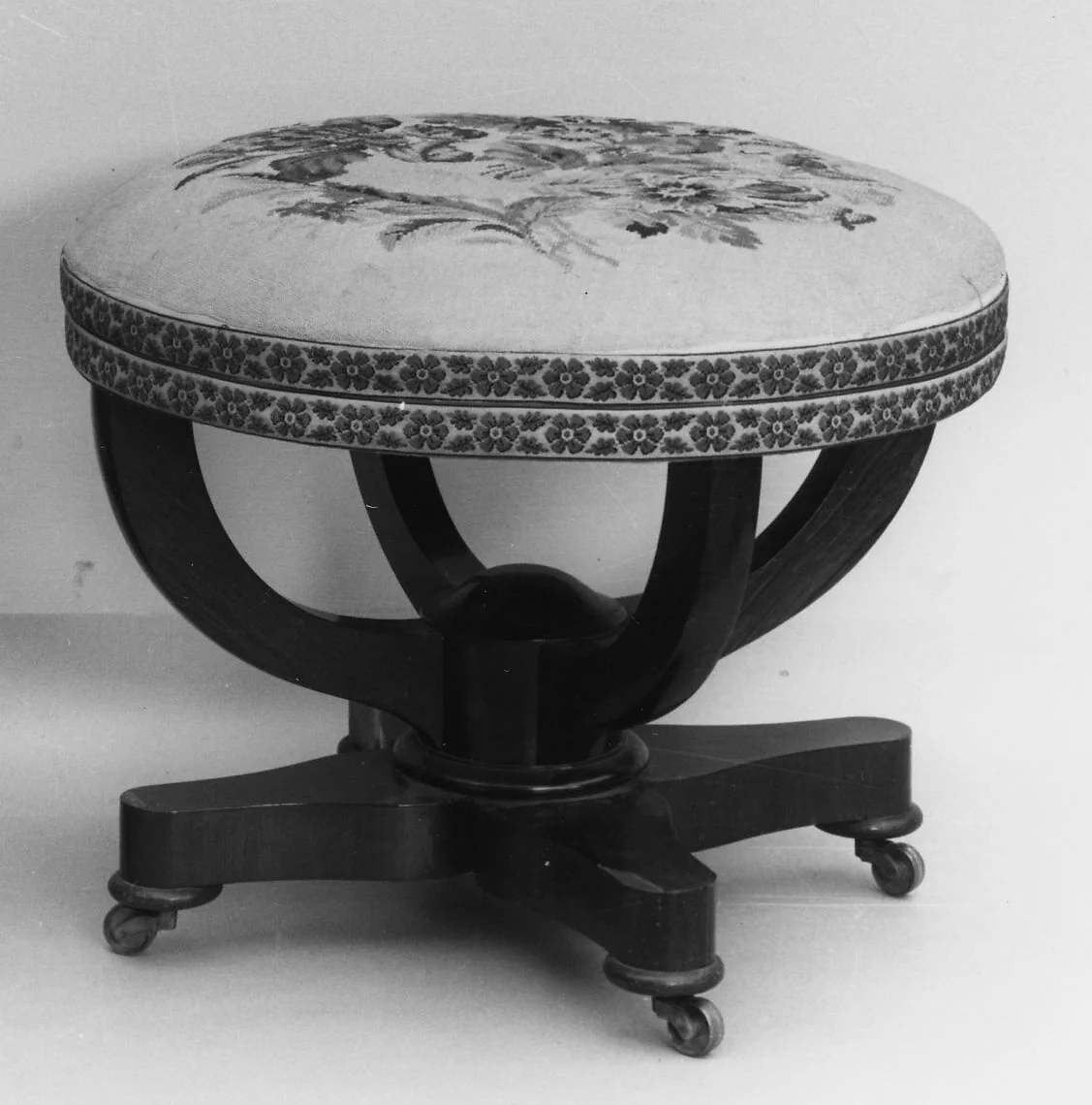 Monochrome photograph of an upholstered round stool with curved wooden supports and a four-footed base on castors.