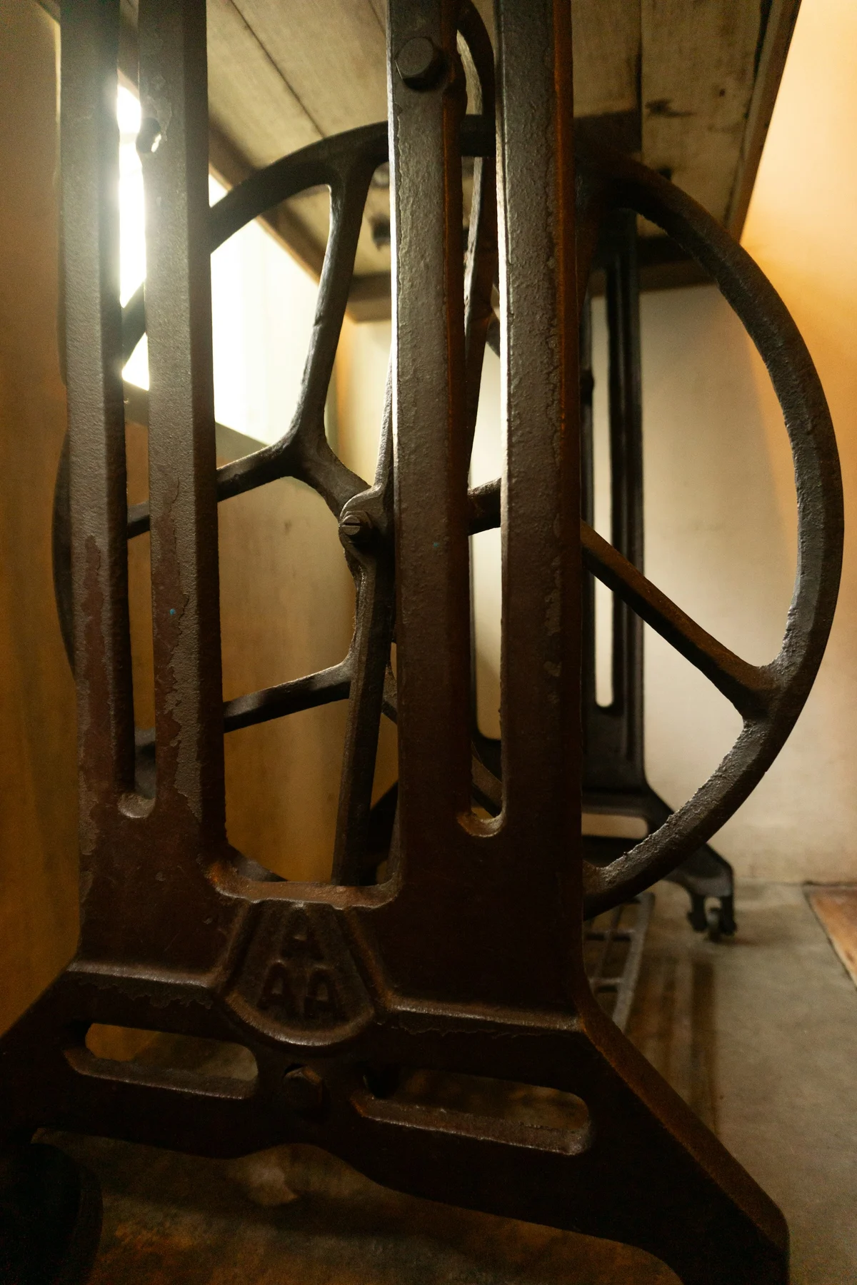 Close view of a metal treadle sewing-machine stand, showing a large wheel, vertical frame and foot pedal below a wooden top.