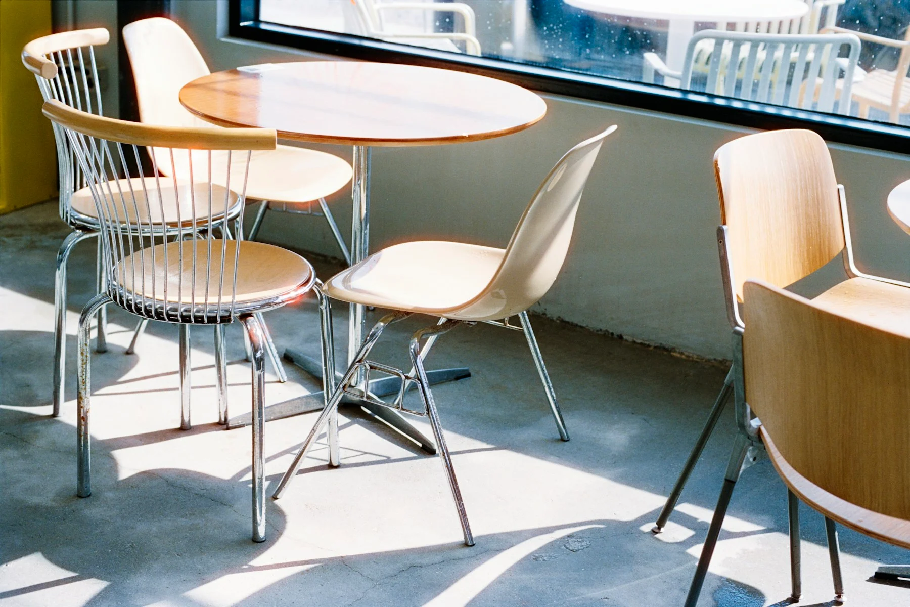Mixed wooden and metal chairs around tables beside a window, with direct sunlight crossing several seats and the floor.