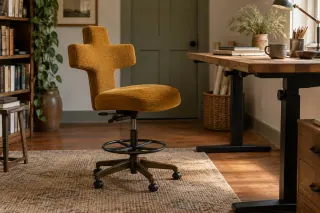 An upholstered saddle chair with a shaped back, foot ring and castor base stands beside a raised desk.