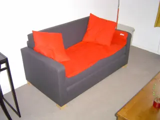Black two-seat sofa with an orange throw and cushions in a living-room corner.