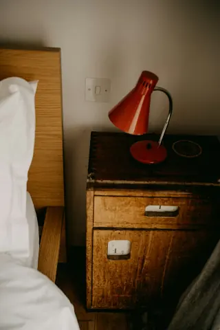 Small wooden drawer cabinet with a red reading lamp beside a bed and wooden headboard.