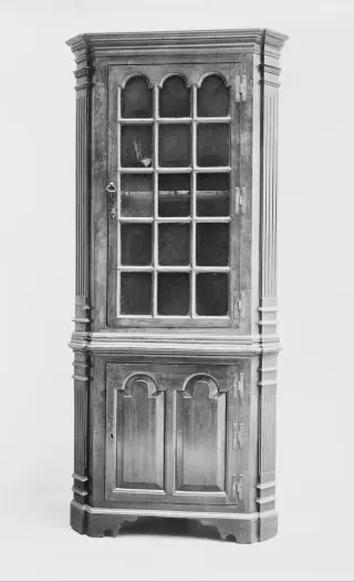 Black-and-white museum photograph of a tall corner cupboard with projecting cornice, glazed upper door and panelled lower door.