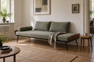 A low black-framed daybed with wooden ends, a green mattress and loose cushions sits against a pale wall.