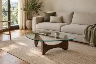 A clear asymmetric glass coffee table with two curved timber base elements stands in front of a pale sofa.