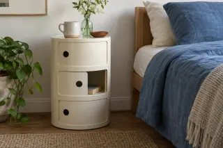 A cream round storage unit with sliding doors stands beside a bed; one compartment holds a paperback.