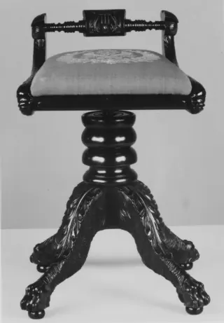 Black-and-white museum photograph of a piano stool with an upholstered seat, small back, central pedestal and four carved legs.