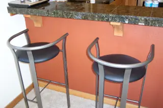 Two tall stools with curved backs, round seats and foot rails standing in front of a kitchen counter with a projecting top and visible underside.