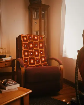 Brown upholstered recliner with curved wooden arms and a crocheted throw, pictured upright beside a table.