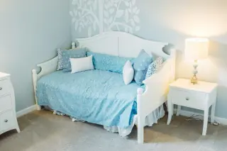 White painted daybed with raised ends, a rear panel, blue bedding and loose back cushions.