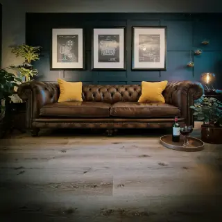 Brown Chesterfield-style sofa with broad rolled arms, deep-buttoned back, two seat cushions and yellow scatter cushions.