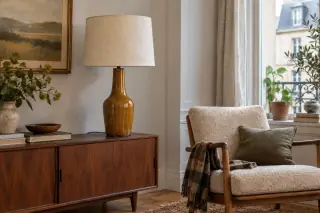A tall honey-brown ceramic lamp with a cream shade stands beside an armchair on a wooden cabinet.