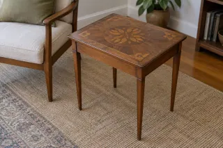 A modest square wooden side table with a patterned decorative top beside an armchair.