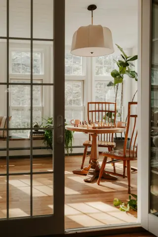 Sunlit glazed room with a small wooden pedestal table, wooden chairs, plants and heating along the window wall.
