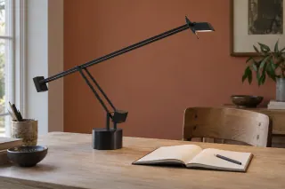 A black counterbalanced task lamp reaches across a wooden desk towards an open notebook.