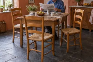 Wooden ladder-back chairs with woven seats around a kitchen table, with a seated reader at the far end.