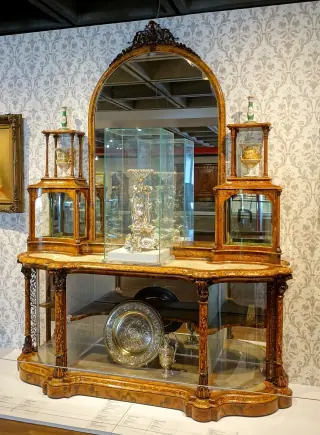 A historic display cabinet with curved glass compartments, multiple shelves, a tall central mirror and a wooden base.