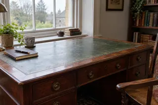 A wooden desk with a green writing inset stands by a window with a notebook and pen.