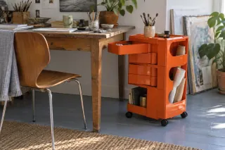 An orange plastic storage trolley with a pivoting tray open stands beside an artist’s desk.