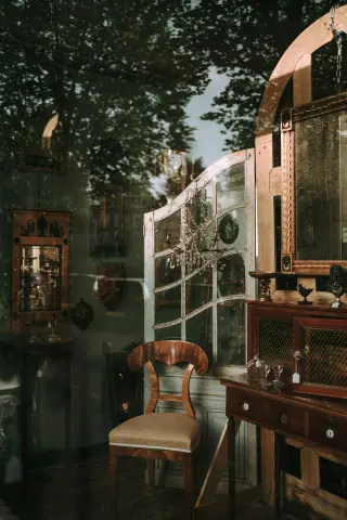 Antique shop window showing a wooden chair, cabinet and glass-fronted display furniture, with reflections of trees.