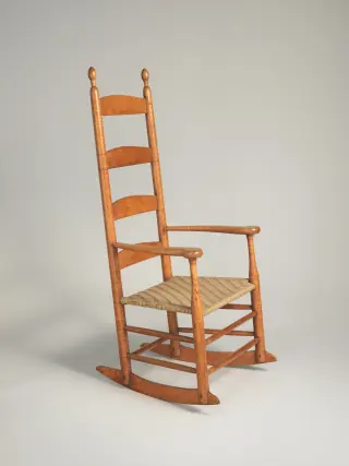 A maple Shaker rocking chair with a tall slatted back, woven seat, arms and two long curved wooden runners, photographed from the front corner.