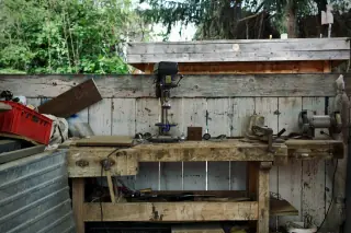 Worn wooden workshop bench with a front vice, lower stretcher and metalworking tools on its top.