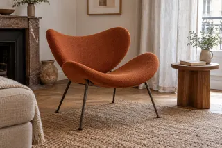 An orange upholstered lounge chair with two separate curved shells and four slim legs beside a wooden table.