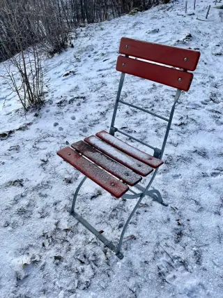 A folding bistro chair with red wooden seat and back slats on a narrow crossed metal frame, standing on snowy ground.