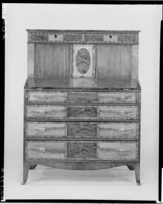Black-and-white photograph of a desk with two vertically slatted shutters beside a central door, above a writing surface and chest of drawers.
