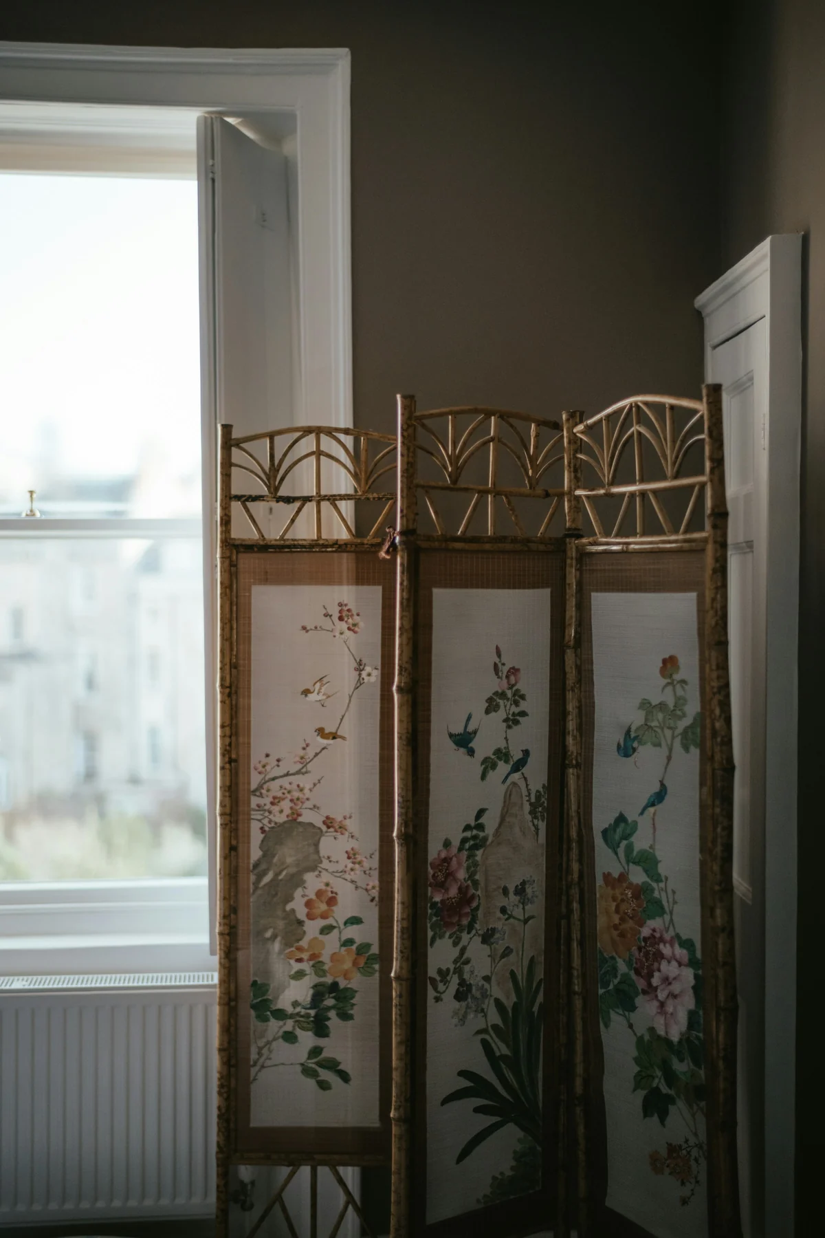 Three-leaf folding screen with bird and flower panels and open geometric upper sections, standing near a window.