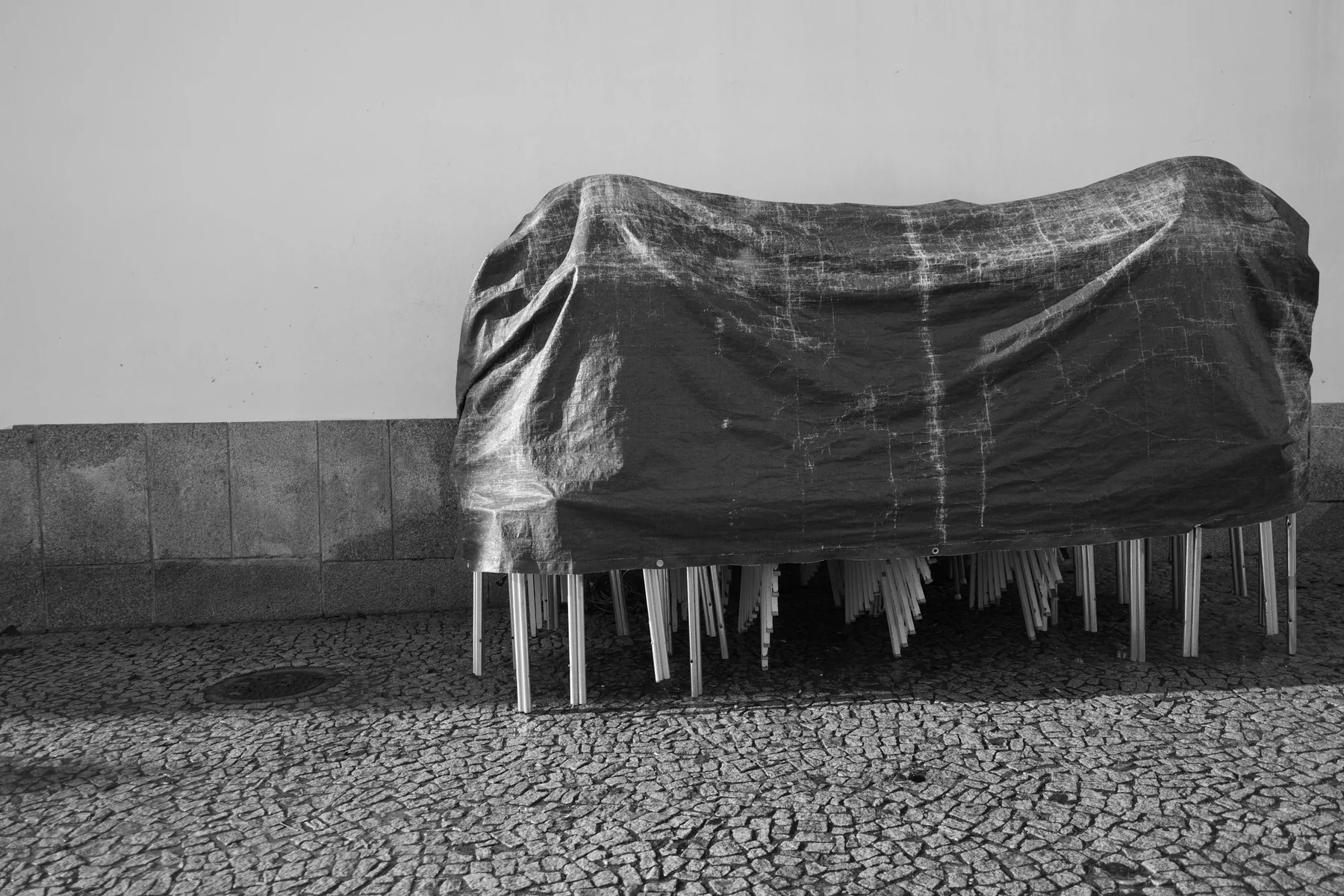 A group of furniture beneath a tied cover outdoors, with chair legs exposed over a cobbled pavement.