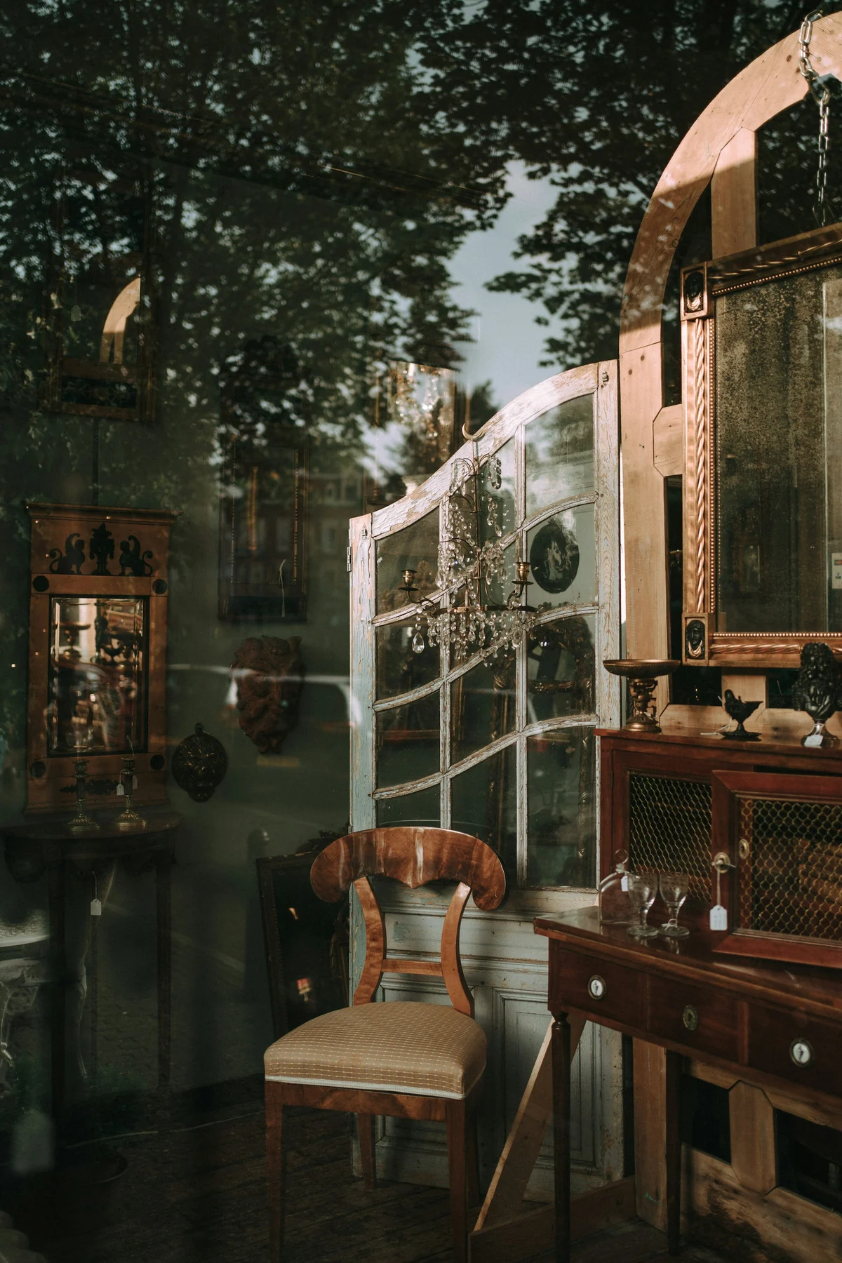 Antique shop window showing a wooden chair, cabinet and glass-fronted display furniture, with reflections of trees.
