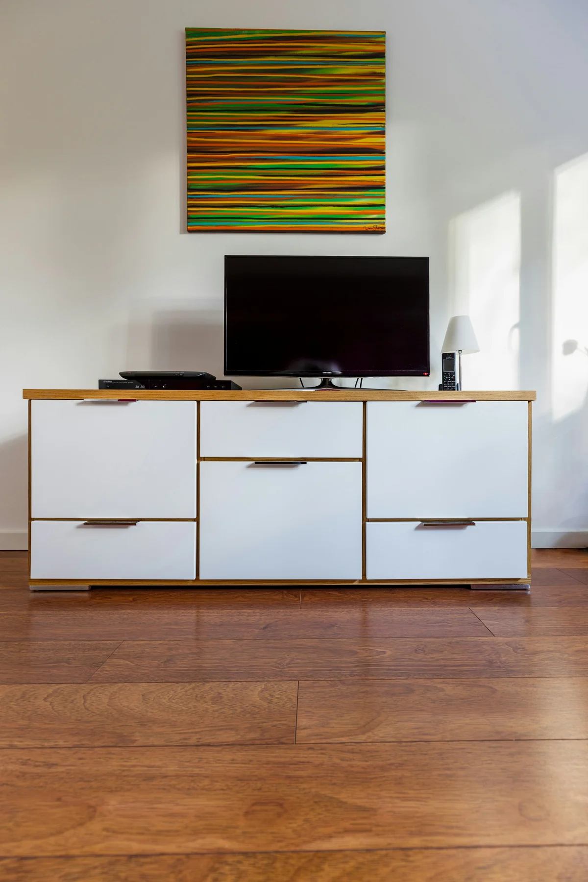 Television on a contemporary cabinet with white drawer and door fronts, a wooden top and framed art above.