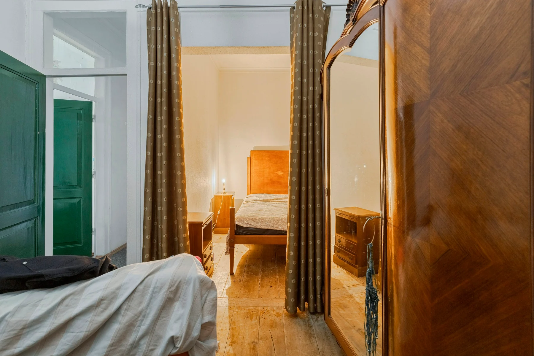 Bedroom viewed past a mirrored wooden wardrobe, with the bed edge, mirror reflection and open doorway visible.